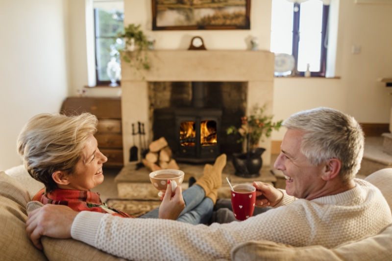 How Do I Keep My Heat During Extreme Cold Weather? Mature couple sat in front of a fire drinking hot chocolate from mugs.