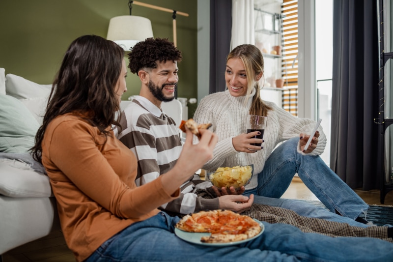 Group of friends having a joyful weekend. They are sitting on the floor, eating, drinking and talking.