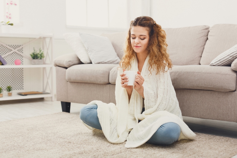 Leaky Ducts and You. Dreaming redhead woman having rest. Wrapped up in white blanket and drinking coffee in warm atmosphere, sitting on floor at home, copy space.