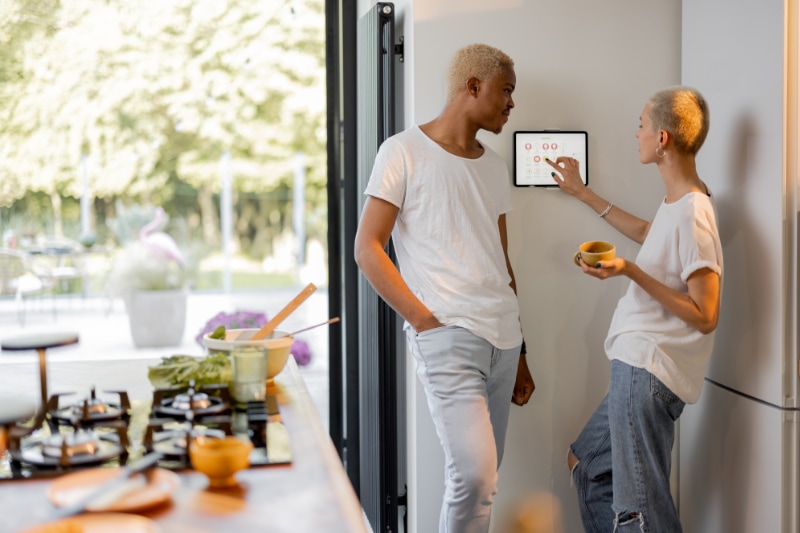 Thermostat 101 Basics. European girl choosing temperature on thermostat of smart home system while her black boyfriend looking at her. Concept of modern technologies in domestic lifestyle. Interior of kitchen.