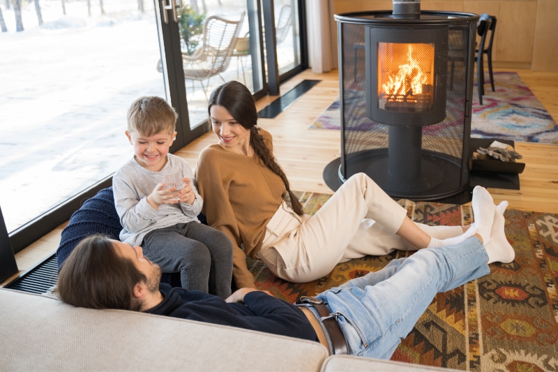 Gas Fireplaces - Staying Safe. Caring mother. Full length view of the nice dark haired woman smiling while sitting at the carpet with her husband and child. Family warming near the fireplace.