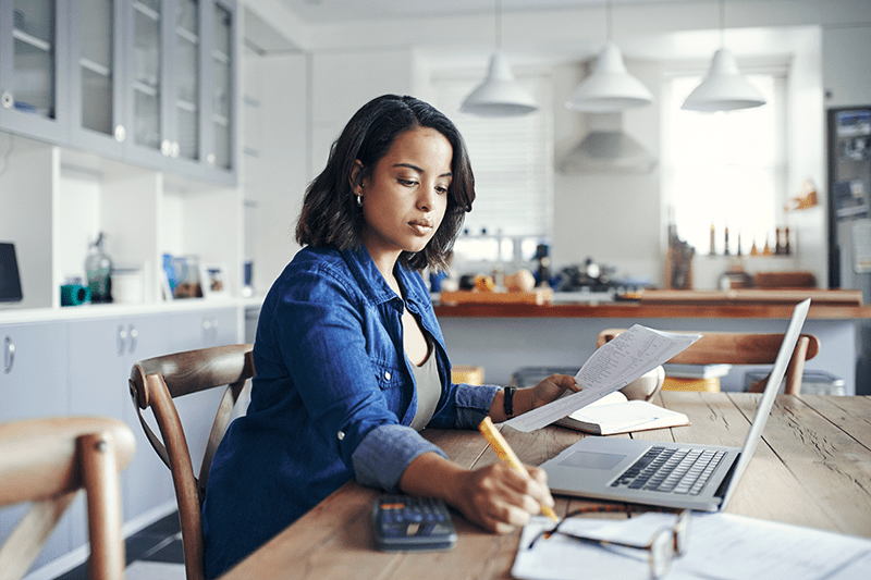 How Can I Lower My Heating Bill This Winter? Image of woman working on computer and pad of paper at kitchen table.